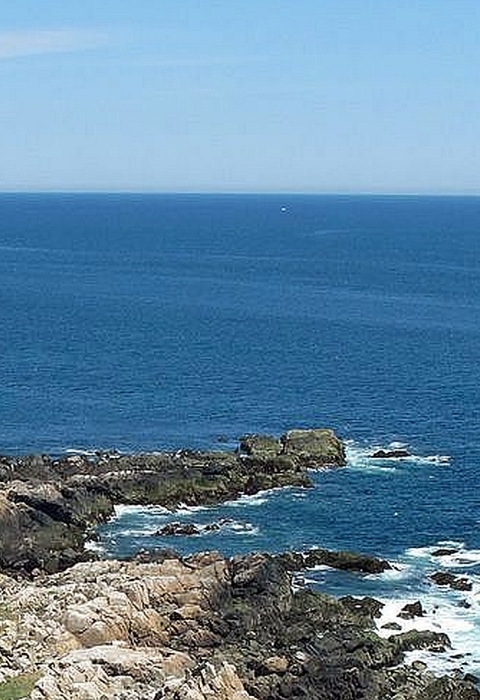 A tall, slender stone lighthouse on a rocky coast of the deep blue ocean