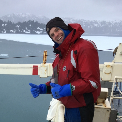 Man wearing orange float coat stands on ship's deck with icy waters surrounding.