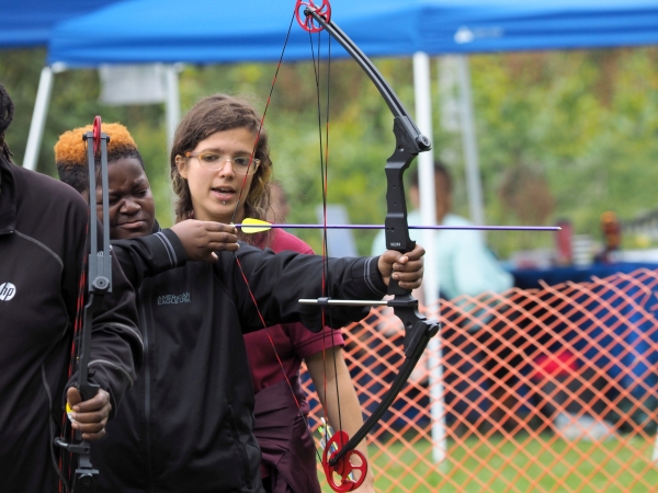 A young adult pulls back on a bow with an arrow drawn, an instructor standing behind him