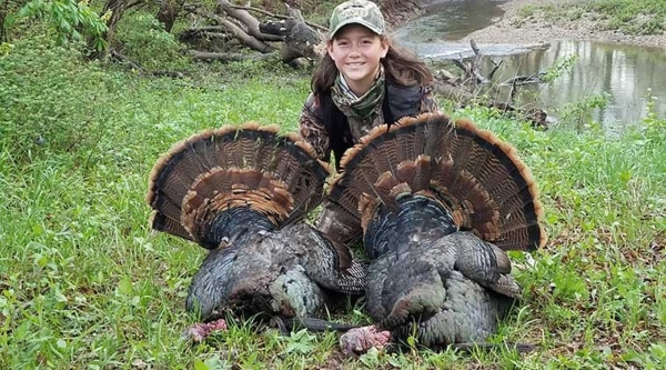 A girl in camouflage crouches on the ground with two large turkeys