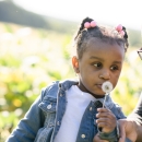 A young girl blows on a puff of dandelion seeds that she picked from the side of the road. She is with a boy, probably her older brother, enjoying the sunshine at the edge of a field. 