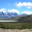 Mountain range with snow-capped peaks in background, and body of water, grass-covered hills, and field in foreground